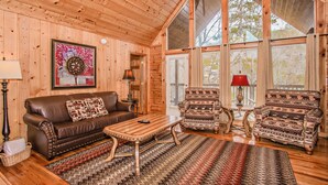 A cozy log cabin interior with a brown leather sofa, patterned armchairs, a wooden coffee table, and a large window offering a view of trees. - 🌲Dancing Bear Den🌄Mountain Views, Resort Pool & Hot Tub (Sevierville)