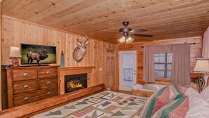 A log cabin room with a fireplace, a bed with a quilt, a mounted deer head, a television, and a ceiling fan. - 🌲Dancing Bear Den🌄Mountain Views, Resort Pool & Hot Tub (Sevierville)