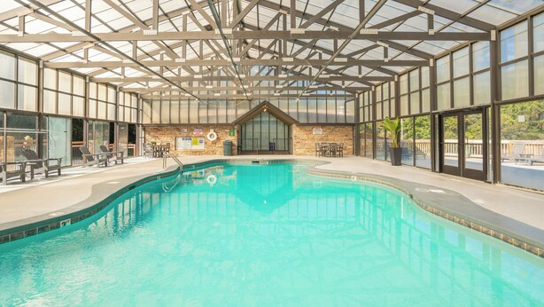 An indoor swimming pool with a glass ceiling, seating areas, and a brick wall with a lifebuoy. - 🌲Dancing Bear Den🌄Mountain Views, Resort Pool & Hot Tub (Sevierville)