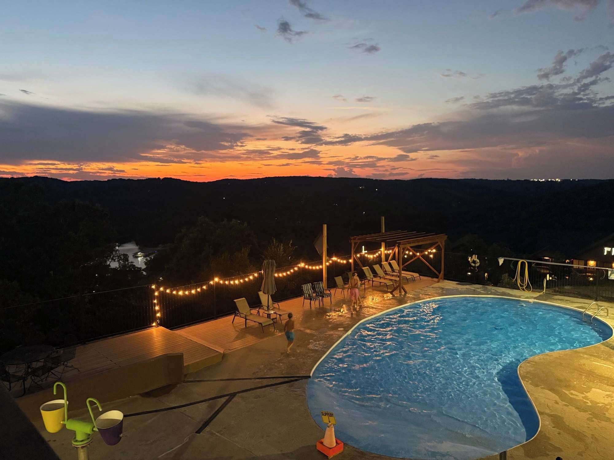 A swimming pool with a view of the sunset, deck chairs, and string lights.