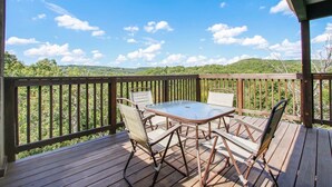 Terraza de madera con una mesa y sillas de vidrio, con vistas a un paisaje boscoso.