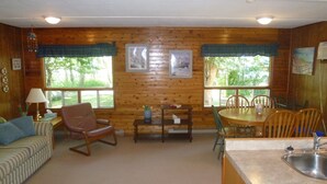 Interior acogedor de una cabaña con una pared de madera, un comedor con mesa y sillas, un sofá, un sillón mecedora y una cocina con fregadero.