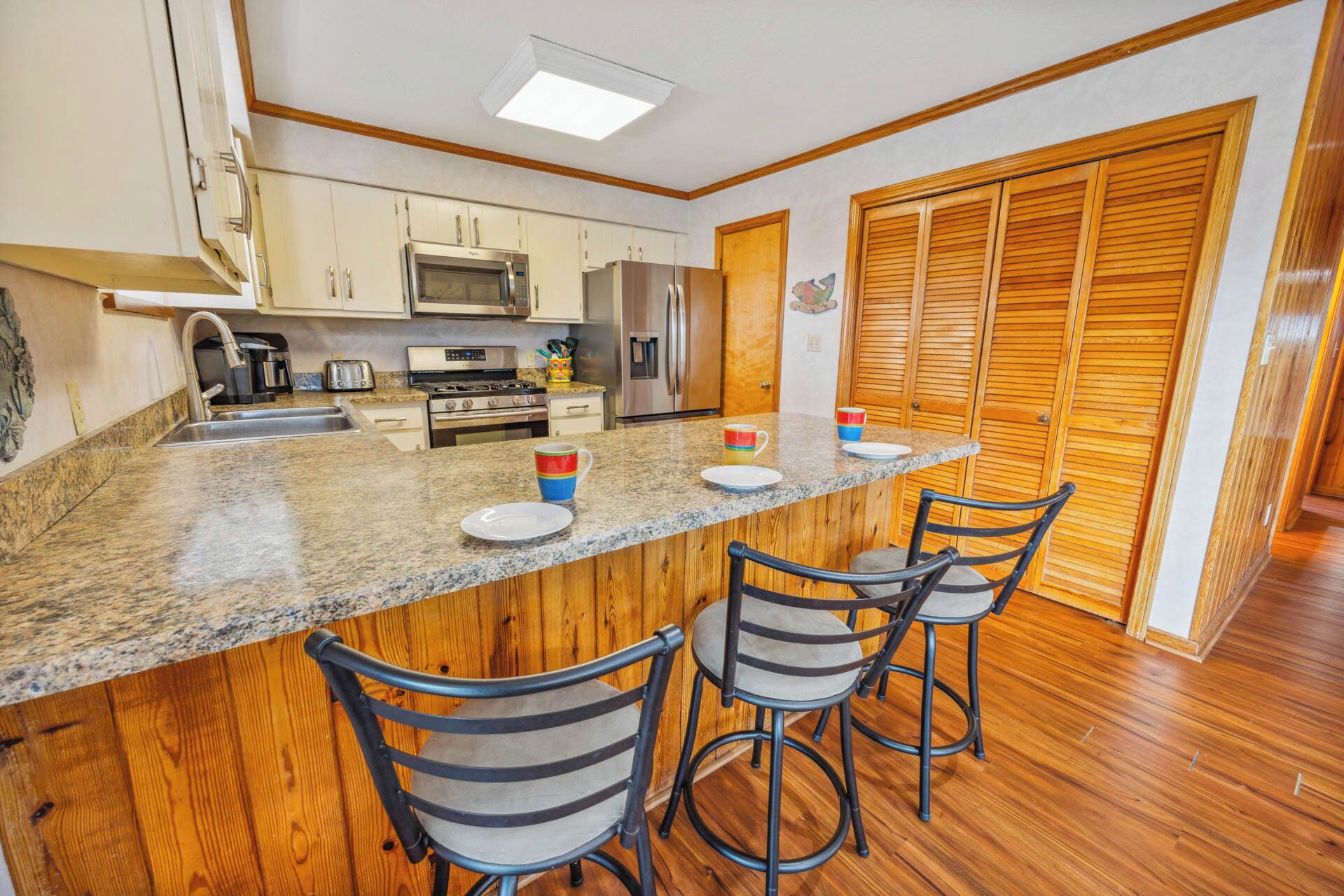 A kitchen with wooden cabinets, a granite countertop, a stainless steel refrigerator, a microwave, and a stove. There are three bar stools and a wooden door with louvered panels.