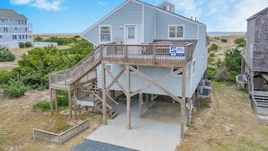 A two-story house with a wooden deck and a sign indicating it is a surfboard rental property. - Oceanfront Avon Home w/ Hot Tub, Foosball, Community Pool and Tennis/Pickleball (Avon)