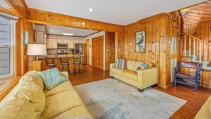 A cozy living room with a sofa and chairs, wooden walls, a kitchen in the background, and a staircase. - Oceanfront Avon Home w/ Hot Tub, Foosball, Community Pool and Tennis/Pickleball (Avon)