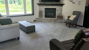A living room with a stone fireplace, a large TV mounted on the wall, and a comfortable seating area with a sofa and chairs. - Close to Aquatic Center, Horicon Marsh, Wedding Venue, Easy Drive to EAA (Waupun)