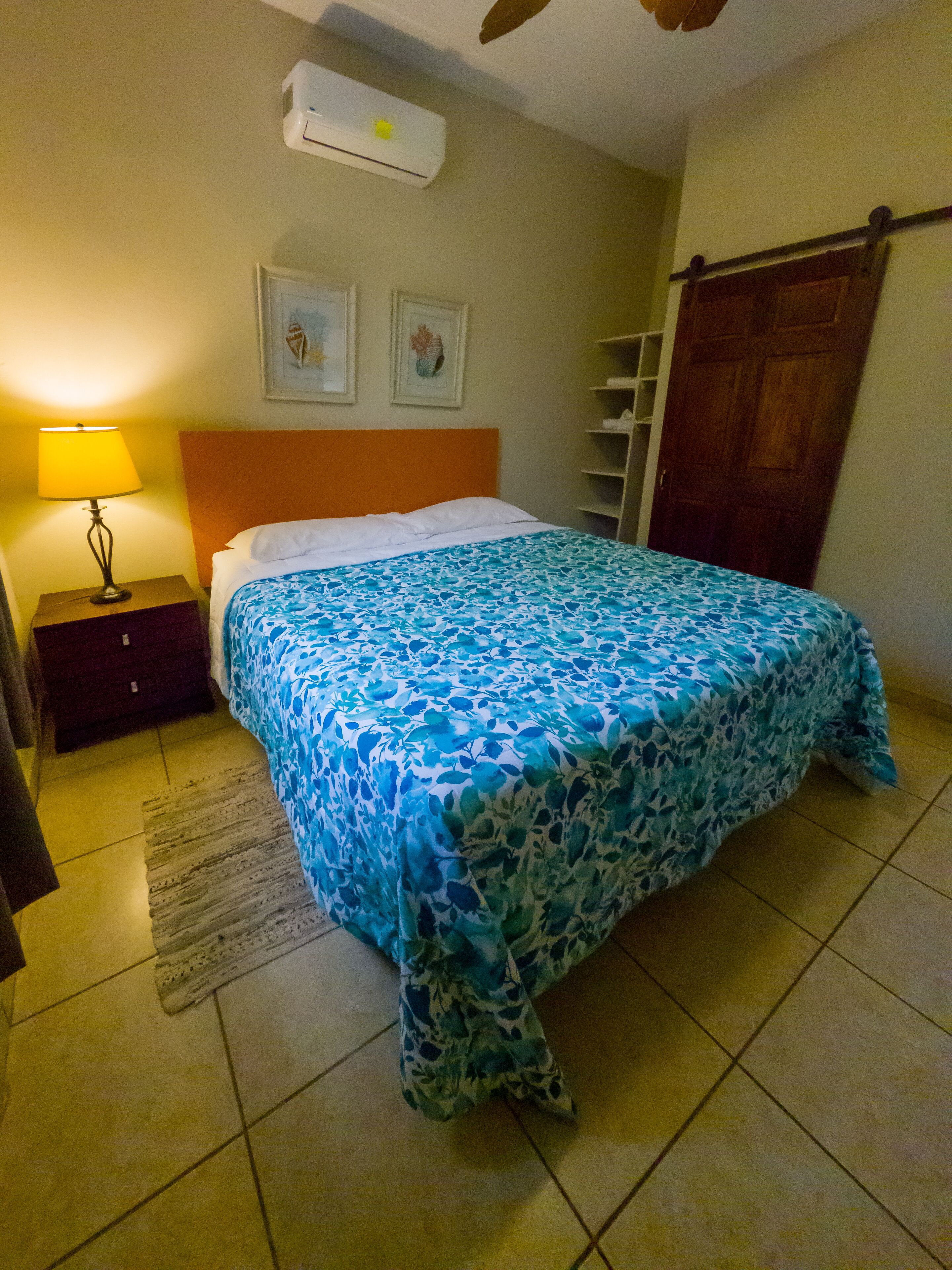 A bedroom with a bed, a nightstand, a lamp, an air conditioner, and a wooden door.