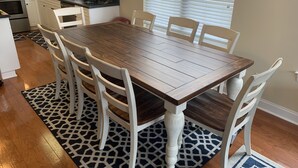 Un coin repas avec une table en bois et des chaises blanches, une cuisine en arrière-plan et un tapis à motifs sur le sol.
