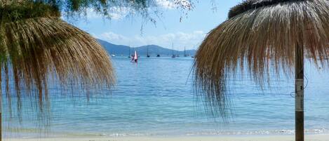 A beach with two thatched umbrellas, a sandy shore, and a calm sea with sailboats.