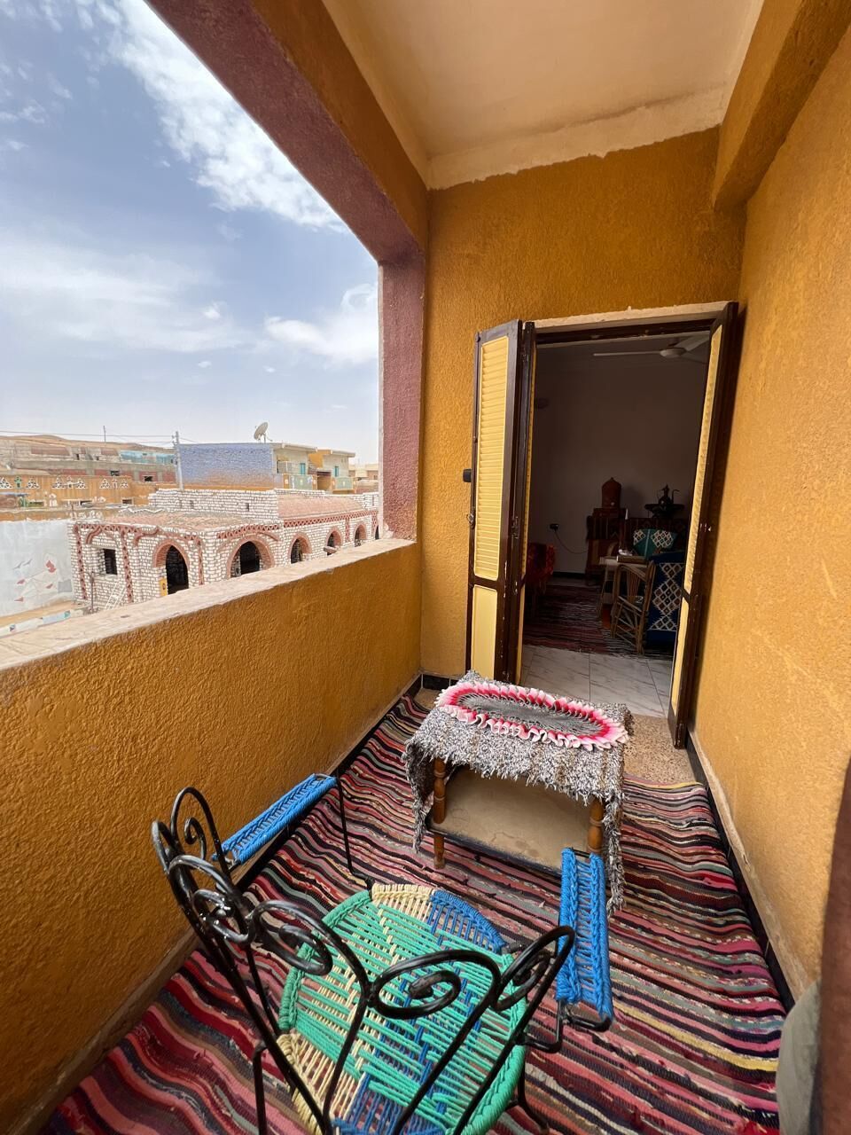 A balcony with a view of buildings, a patterned rug, and a small table with a chair.