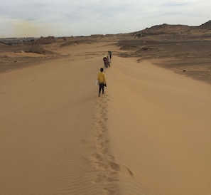 A desert landscape with a group of people walking towards a distant building. - Nubian Kindom Aragheed House (Aswan)