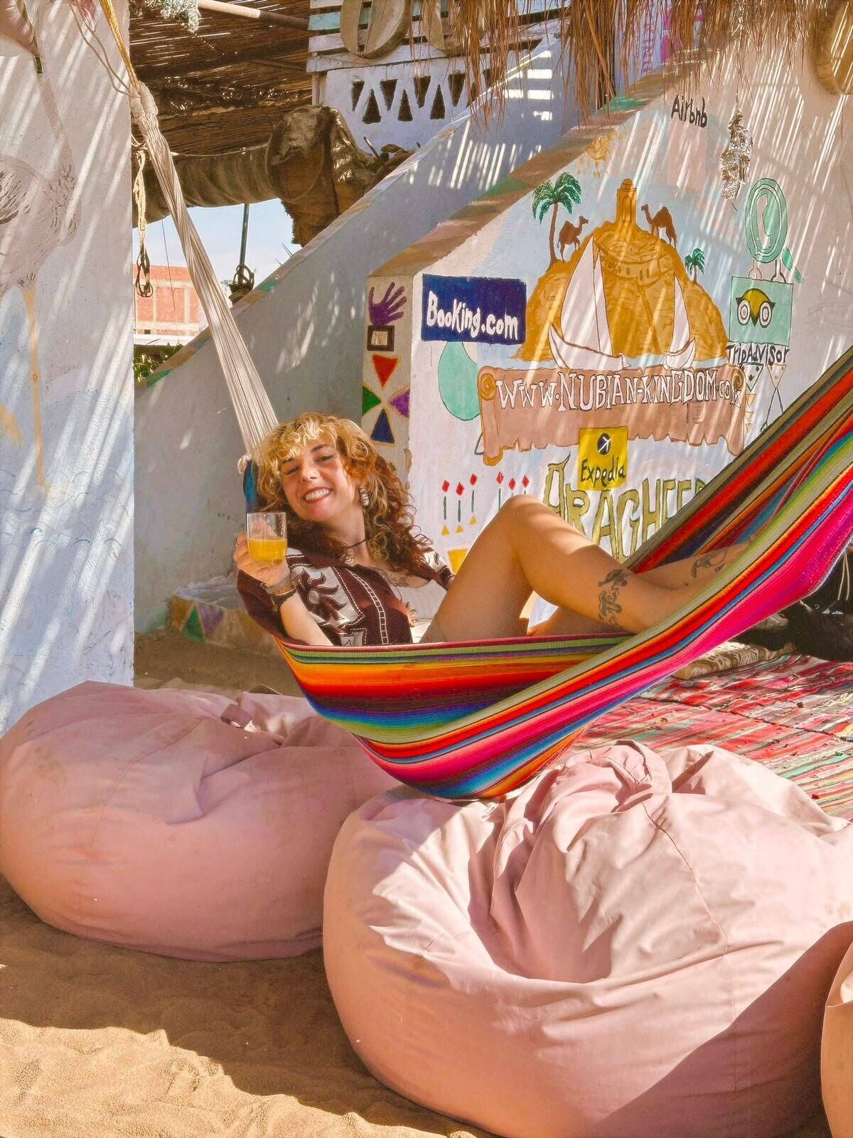 Una persona relajándose en una hamaca con tela a rayas de colores, sentada en una playa con un mural de fondo que muestra varios logotipos y texto relacionado con viajes.