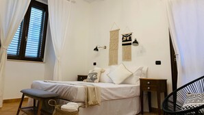 Un dormitorio con cama, mesitas de noche, una silla, una ventana con cortinas e iluminación en la pared.