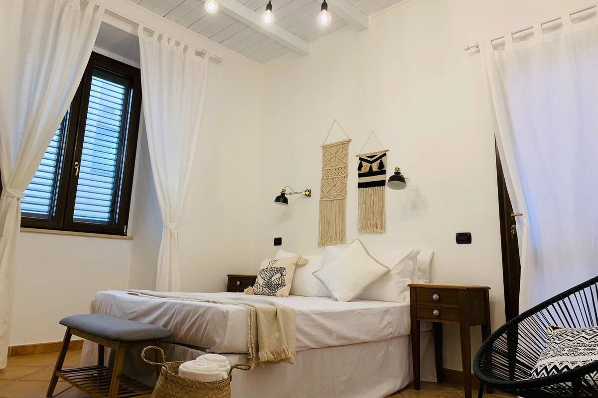 Un dormitorio con cama, mesitas de noche, una silla, una ventana con cortinas e iluminación en la pared.