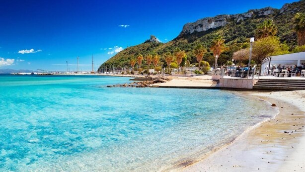 Una playa con agua turquesa cristalina, una colina rocosa al fondo y una zona de estar cubierta con gente.