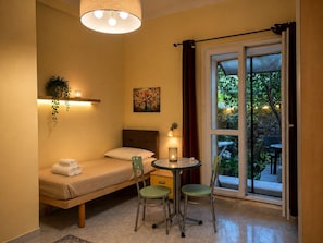 A compact room with a bed, a small table, two chairs, a wall-mounted lamp, a painting, and a window with curtains. - Andrea Apartment.  (Portoscuso)