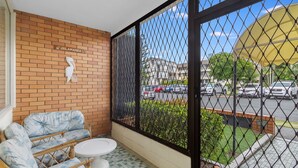 Ein Balkon mit Korbmöbeln, einem weißen Tisch und Blick auf einen Parkplatz sowie Gebäude.
