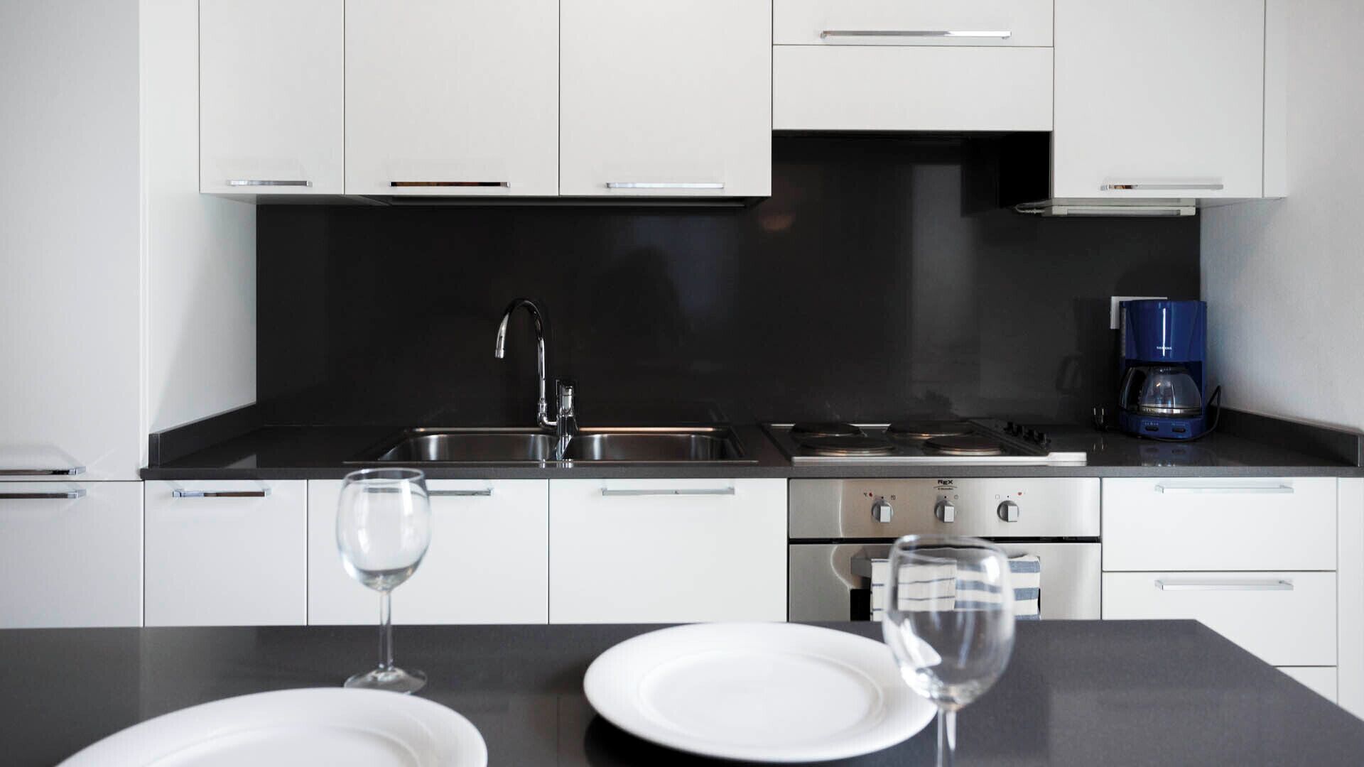 A modern kitchen with white cabinets, a black countertop, a stainless steel sink, a stove, and a coffee maker.