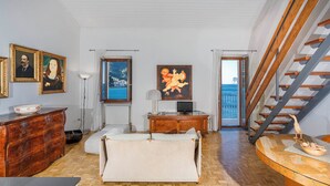 Una habitación con piso de madera, un sofá, un armario de madera, un cuadro, una lámpara y vista al mar por una ventana.