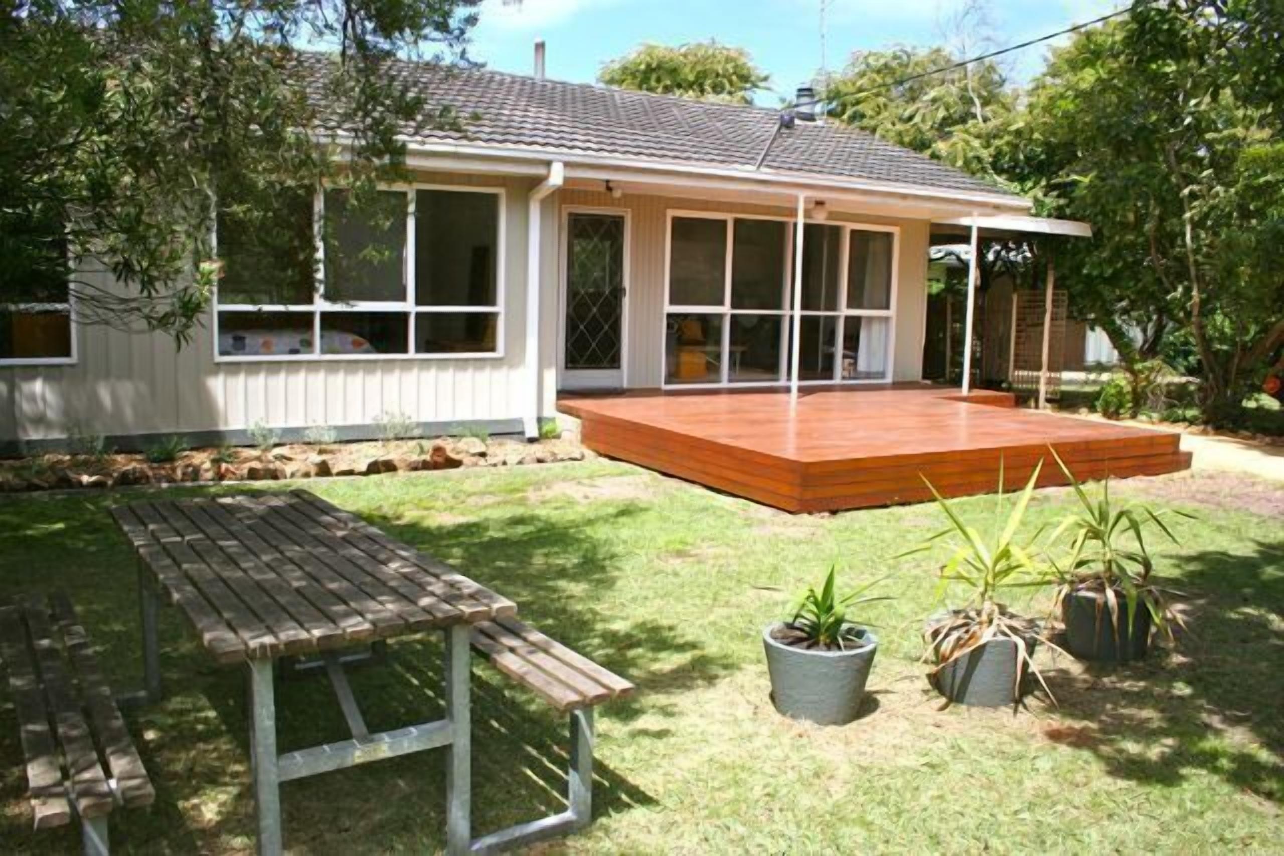 Uma casa com deck de madeira, mesa de pátio e plantas em vasos.