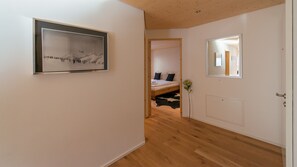 A hallway with a wooden floor, a framed picture on the left, a bed with pillows in the background, and a mirror on the right.