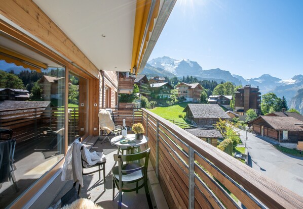 A balcony with a table and chairs, overlooking a mountainous village with traditional houses.