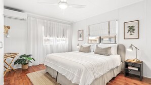 Een slaapkamer met een groot bed, een nachtkastje, een lamp en een raam met dunne gordijnen.