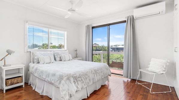 Un dormitorio con cama, mesita de noche, lámpara, ventilador de techo, ventana con persianas, balcón con vistas y aire acondicionado.