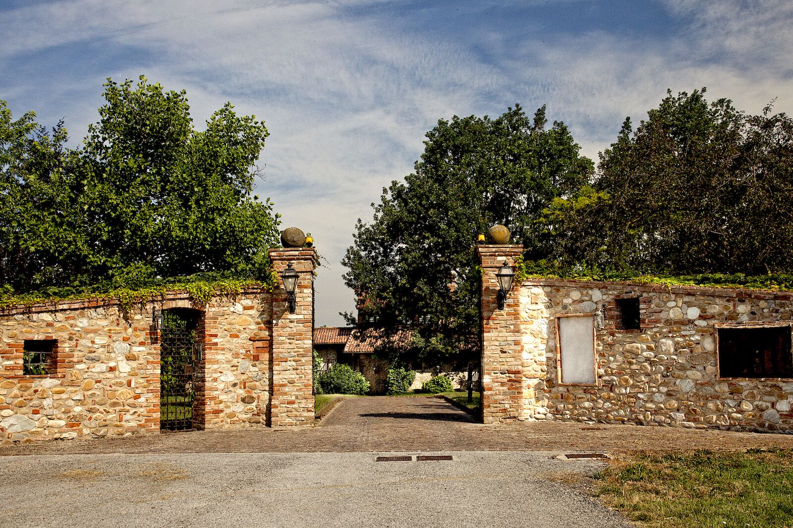 Un portón de piedra con una puerta metálica, que da a un edificio con un muro de piedra y una ventana pequeña.