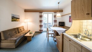 A compact room with a sofa, a small table, a dining area, a kitchen with a sink, and a television.