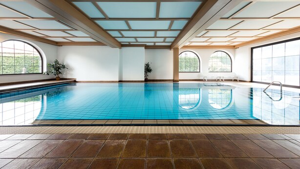 An indoor swimming pool with a tiled floor, wooden ceiling beams, and large windows.