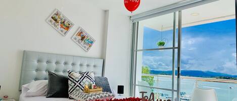 Un dormitorio con una cama grande, un ventanal con vistas al mar y globos rojos con la frase 'Te Amo'.