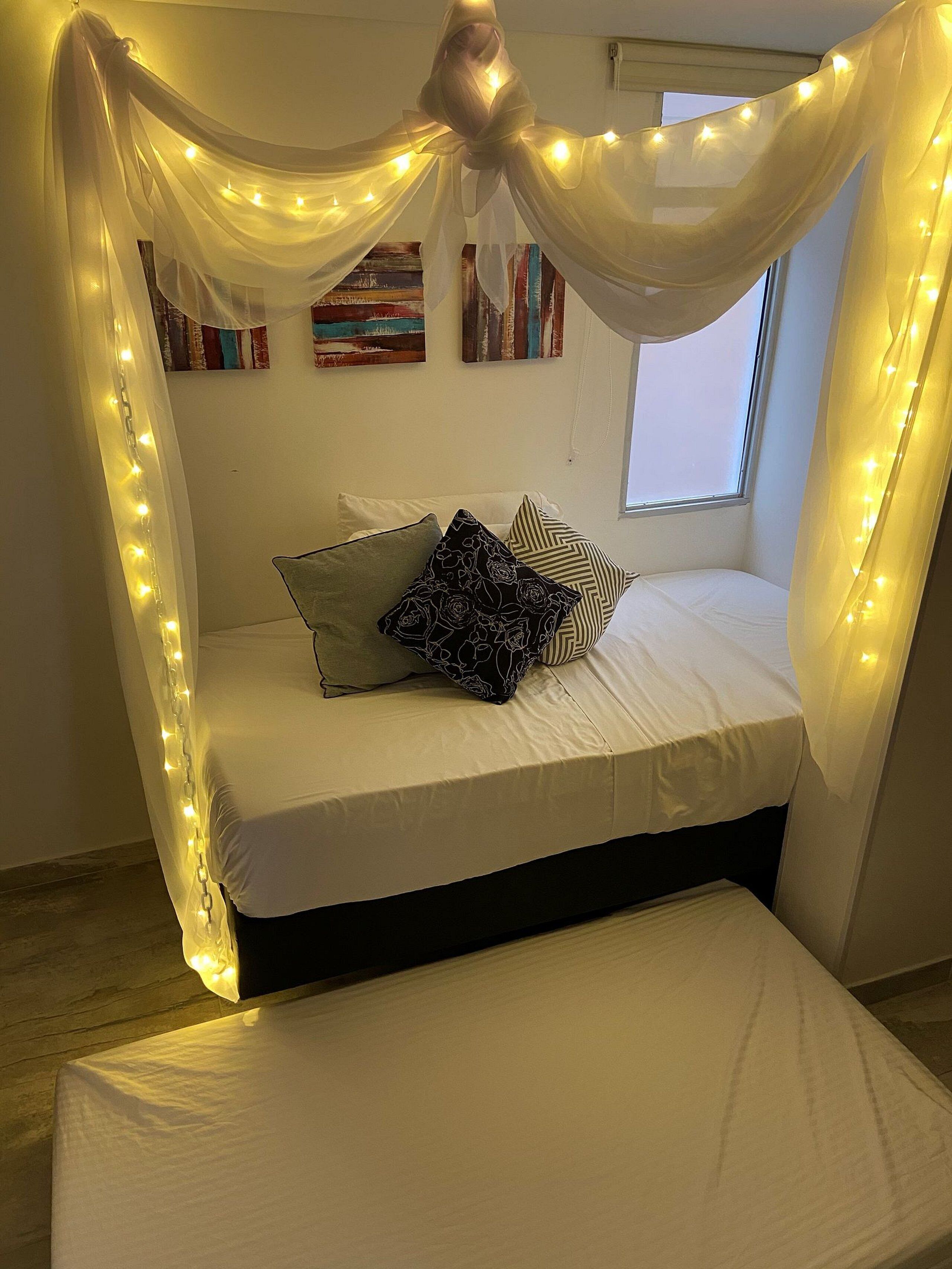 A bed with a canopy and fairy lights, a window, and a painting on the wall.