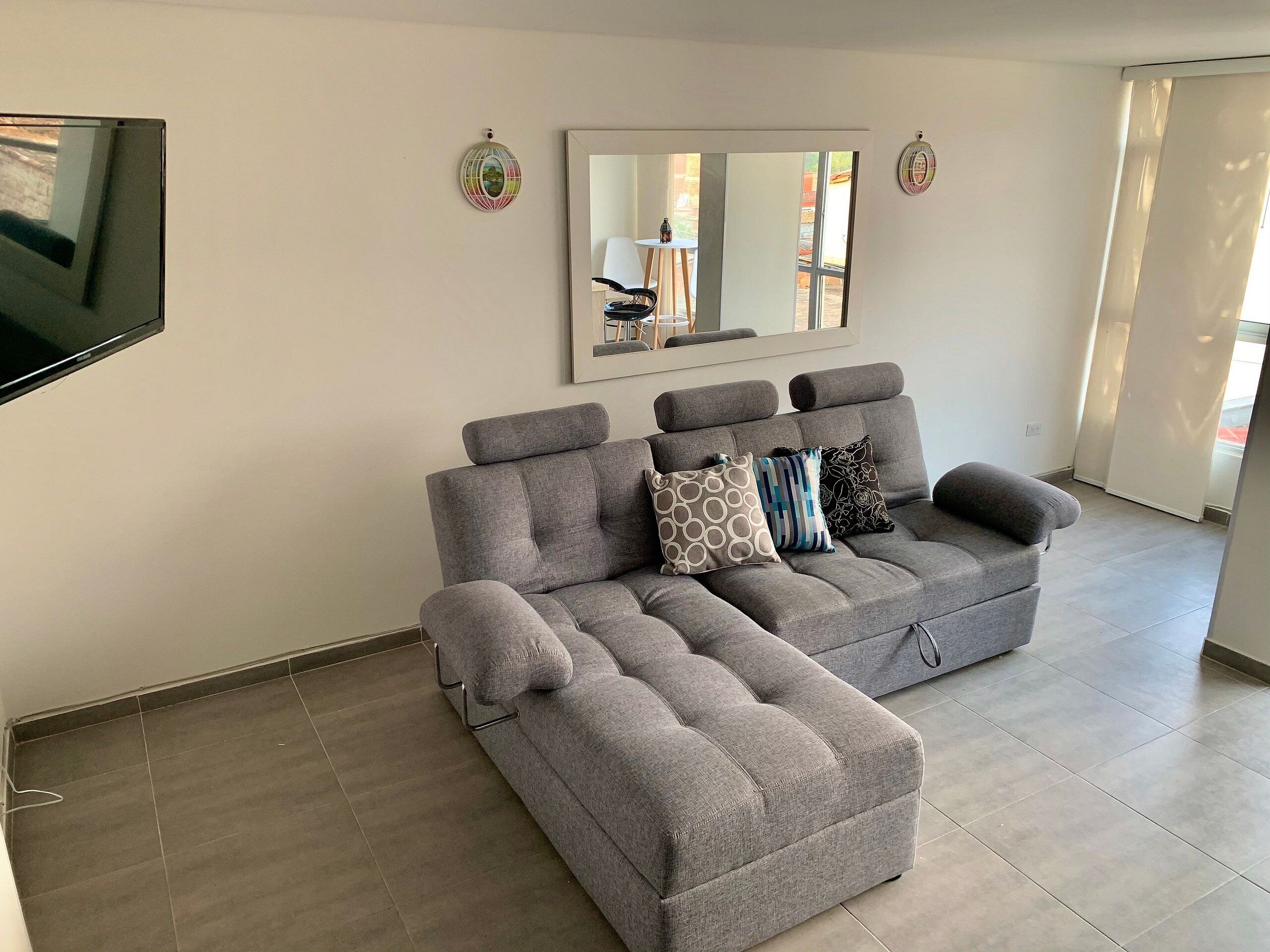 A grey sectional sofa with three cushions, a flat-screen TV mounted on the wall, and a large mirror above the sofa.