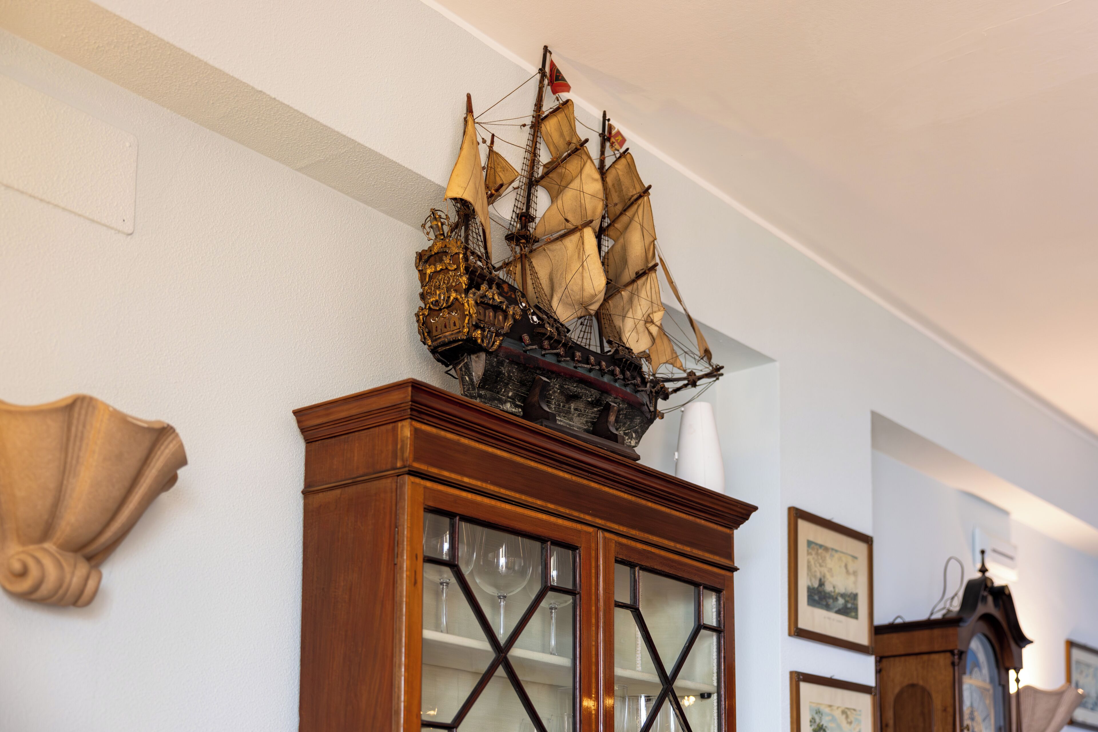 Un mobile in legno con ante in vetro, un modellino di nave sopra e quadri appesi al muro.