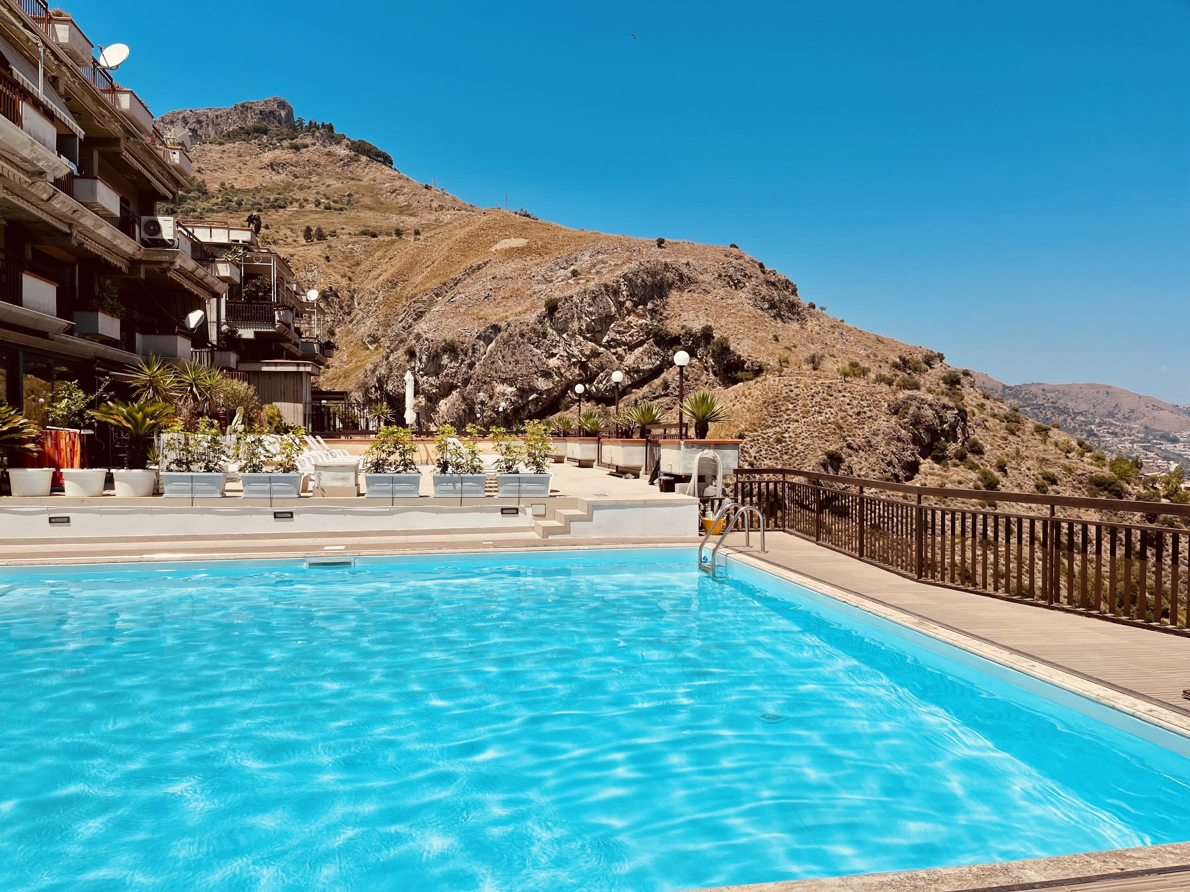 Ein Schwimmbecken mit Holzterrasse und Liegestühlen, eingebettet in eine gebirgige Landschaft.