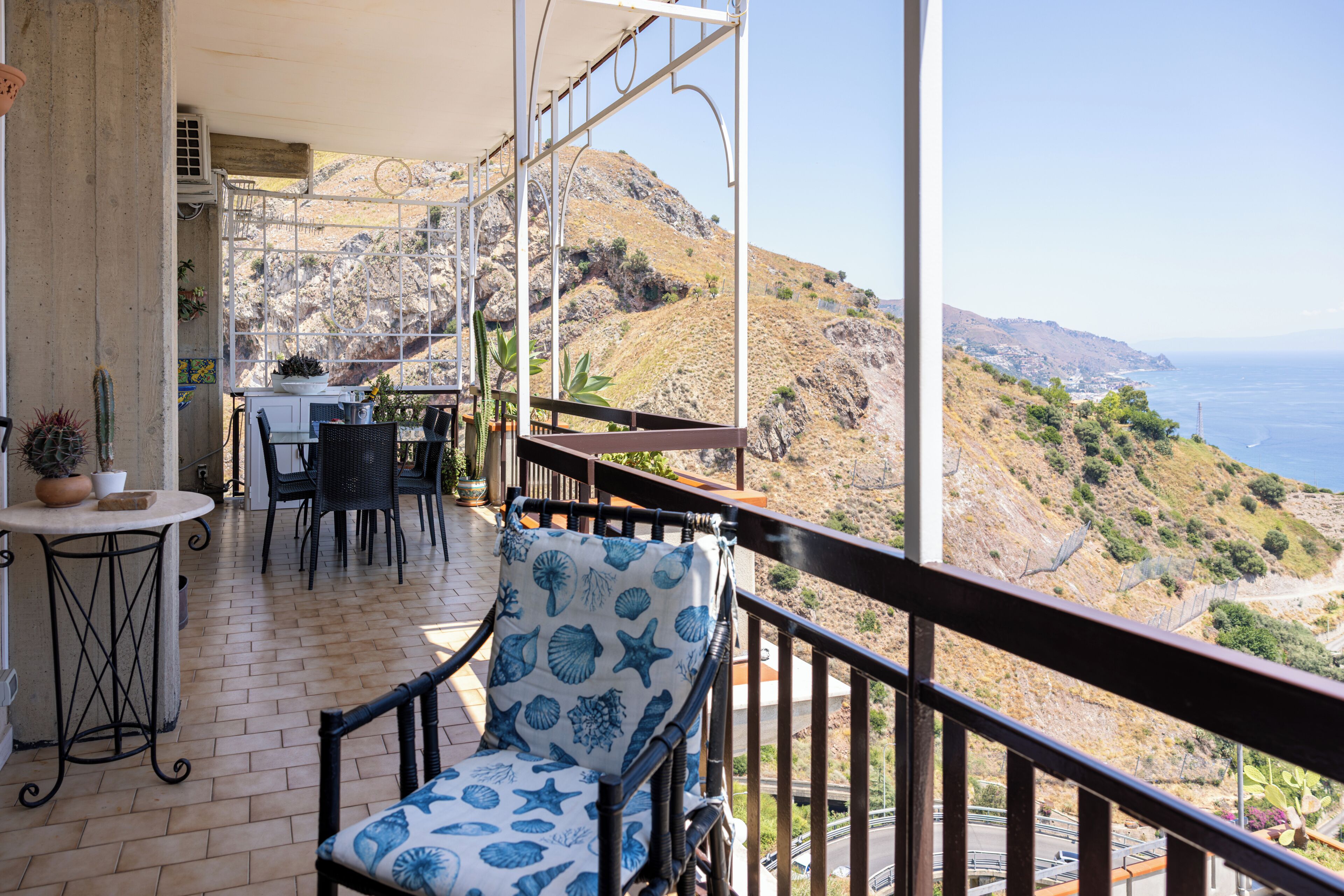 A balcony with a table and chairs, overlooking a mountainous landscape and the sea.