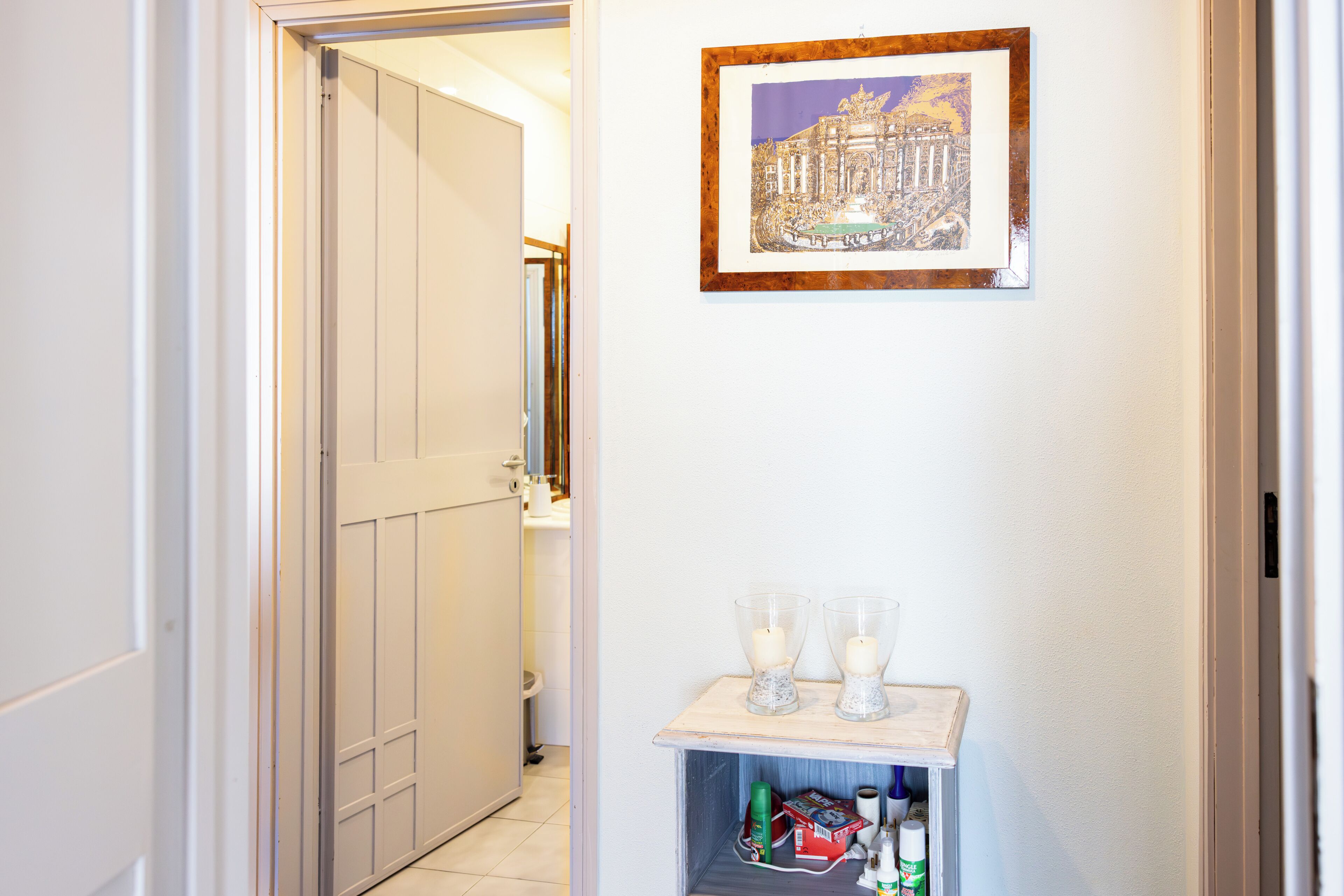 Un corridoio con una porta che conduce al bagno, uno scaffale con candele e un quadro appeso al muro.