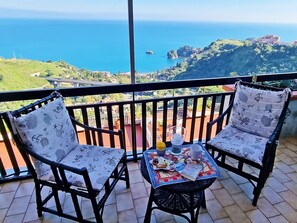 A balcony with two wicker chairs, a small table with a colorful tablecloth, and a view of the ocean and greenery. - A splendid three-room apartment with a pool and breathtaking views. (Taormina)