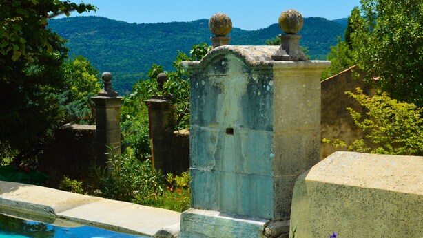 A stone structure with a pool, surrounded by greenery and hills in the background.