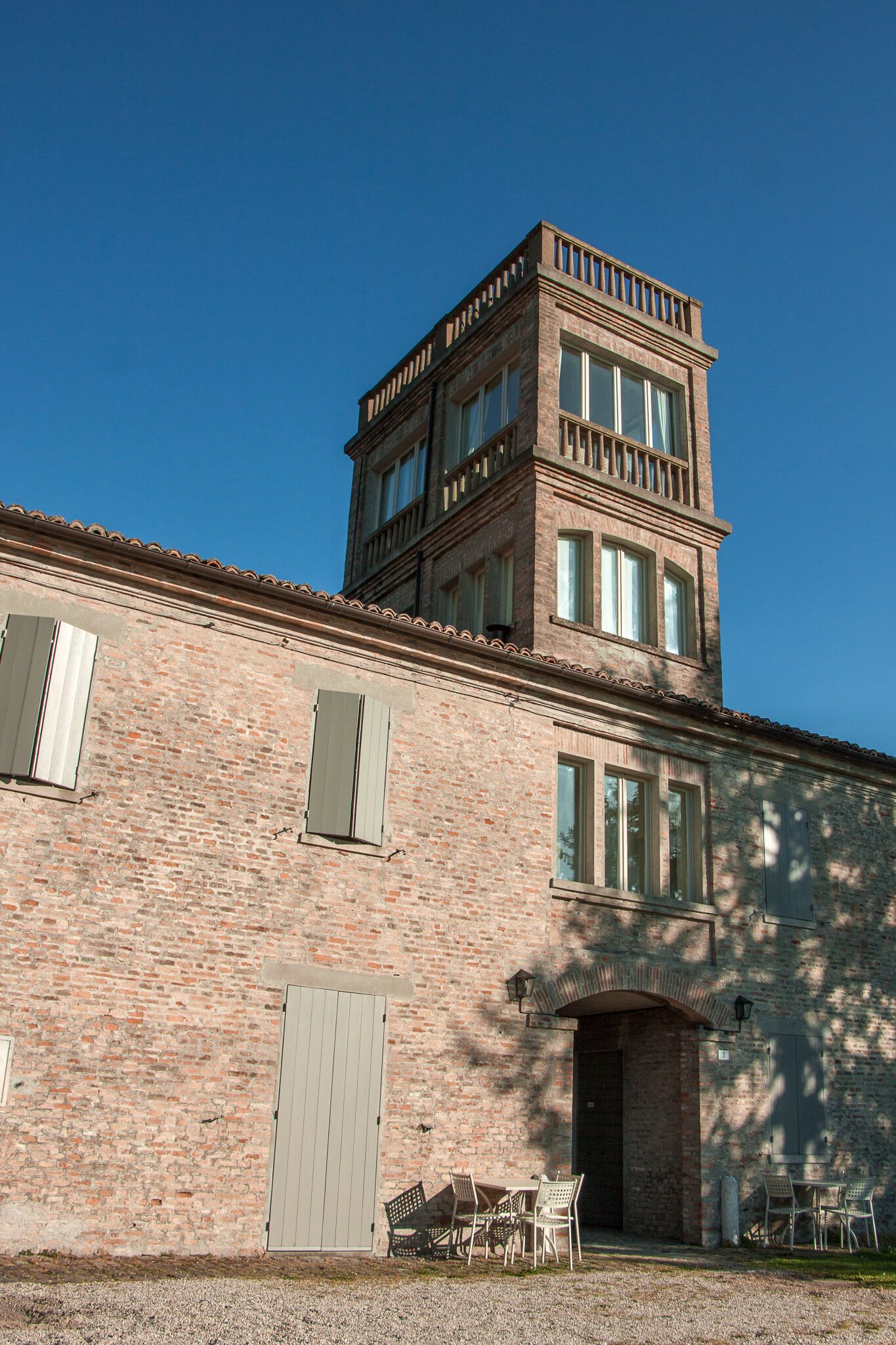 A two-story brick building with a balcony on the upper floor, a door, and a small outdoor seating area with tables and chairs.