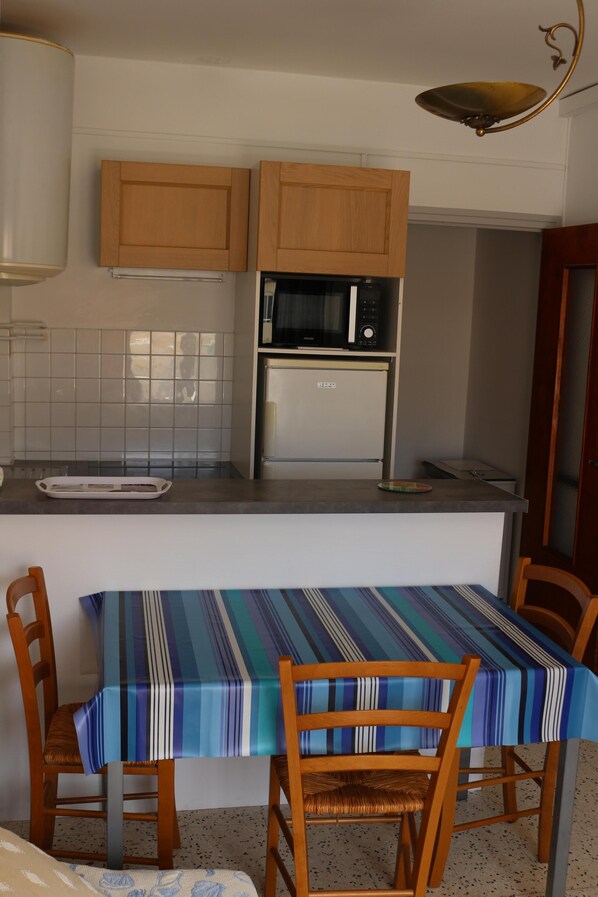 Una cocina compacta con armarios de madera, microondas y refrigerador. Hay una mesa de comedor con mantel a rayas y dos sillas.