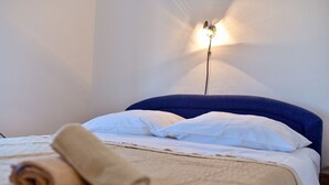 A bed with a blue headboard, white pillows, and a beige blanket rolled at the foot. A wall-mounted light fixture casts a shadow on the wall. - Apartment in Vir with Terrace, Air condition, WIFI (4592-1) (Vir)