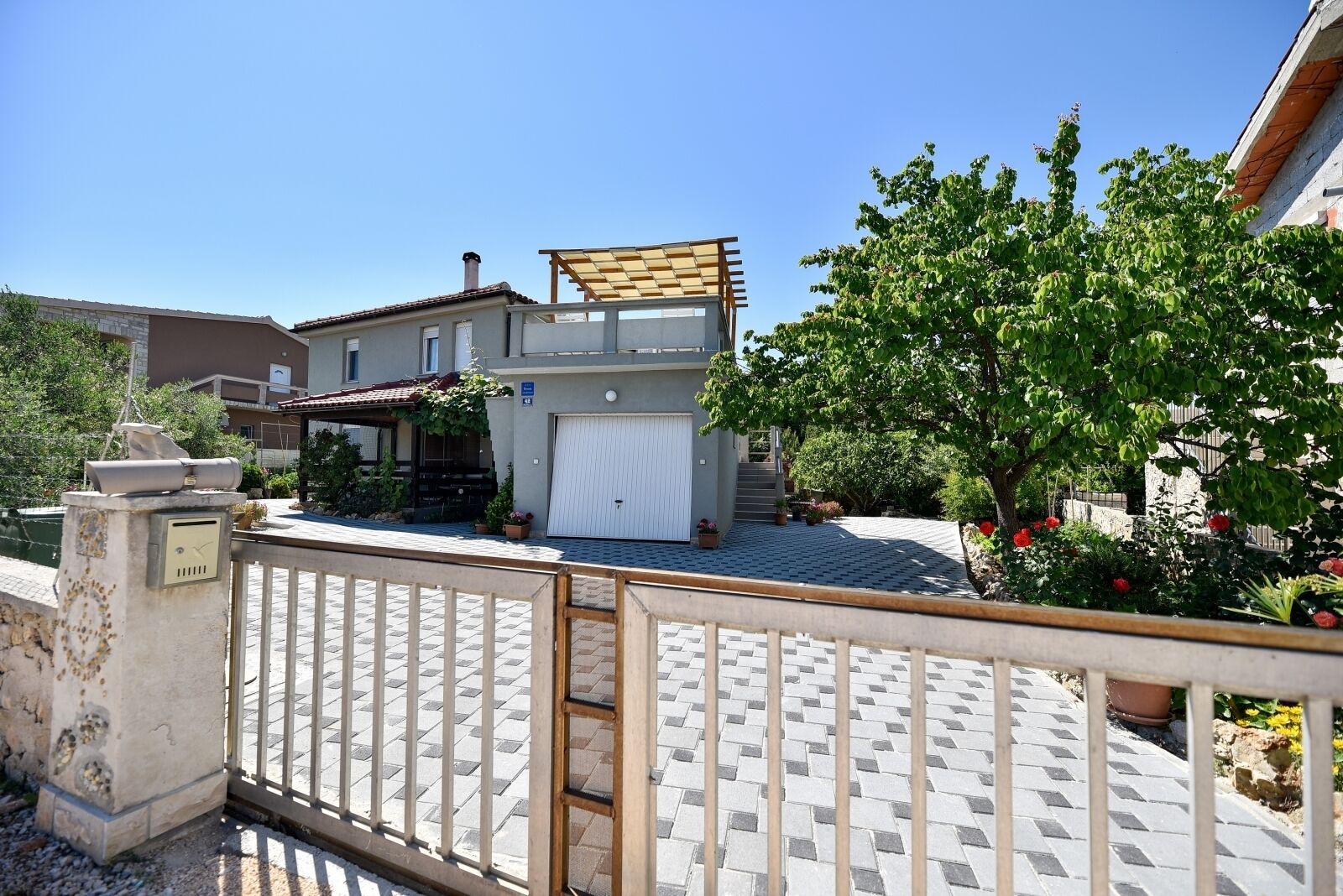 Ein zweistöckiges Haus mit überdachter Terrasse, Garage und Garten mit Kübelpflanzen und Blumen.
