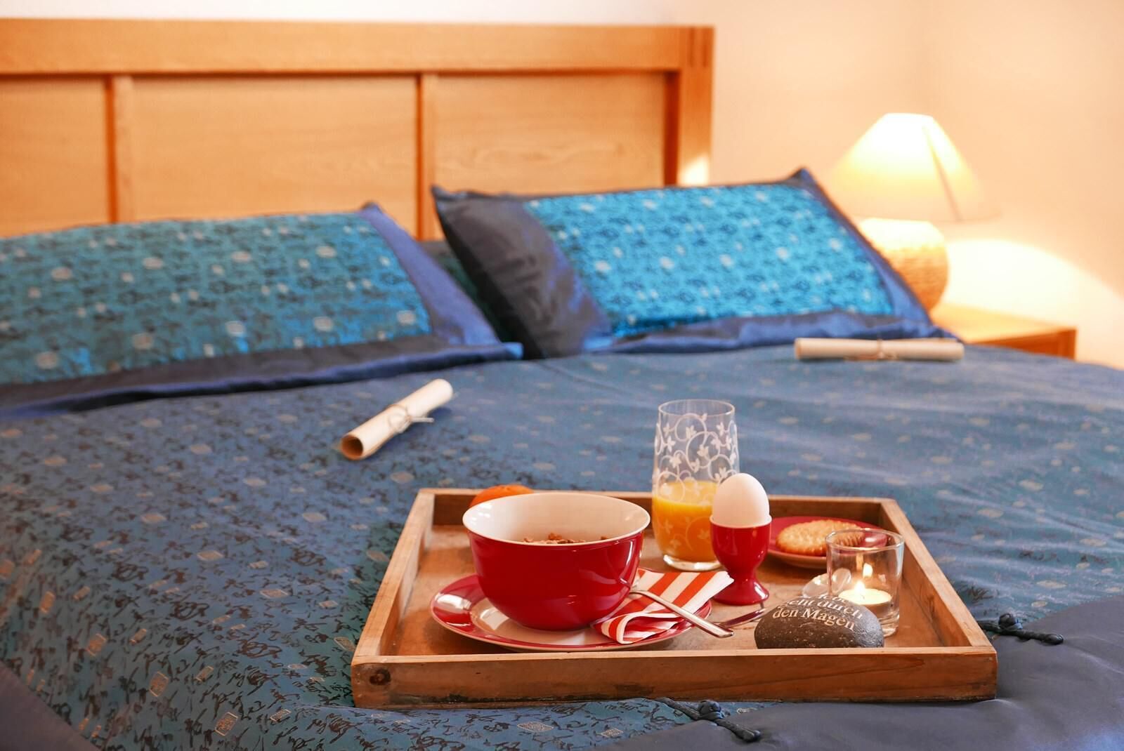 A neatly made breakfast tray on a bed with a blue patterned bedspread, a red bowl, a glass of orange juice, a candle, and a small plate with a cookie.