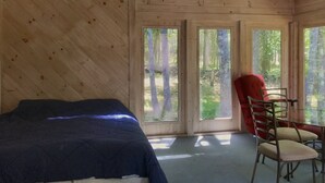 Una habitación con cama, una silla roja y grandes ventanas con vista al bosque.