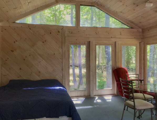 A room with a bed, a red chair, and large windows with a forest view.