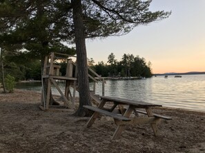 A wooden picnic table and a tree by a calm lake with boats in the distance.
