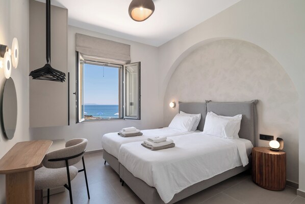 A bedroom with a bed, bedside table, chair, and a view of the sea through an open window.