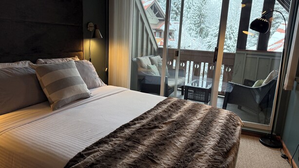 A hotel room with a large bed, a fur throw, a glass door leading to a balcony with outdoor furniture.